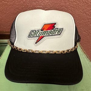 May include: A white and black trucker hat with a gold chain around the brim. The hat has a red, orange, and green embroidered patch with the text "Gatumadre" on it.