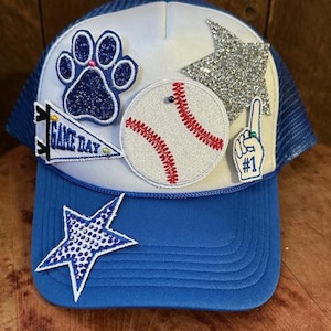 May include: Blue and white baseball cap with a curved brim. The cap features a paw print, baseball, star, and #1 hand patches. The text "GAME DAY" is on a pennant patch. The cap has a mesh back.