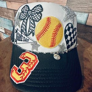 May include: A black and white trucker hat with a white crown and a black bill. The hat has a variety of patches, including a softball, a bow, a heart, and a star. The hat also has a silver chain with a softball charm. The bill of the hat has a red and yellow number 3 patch.