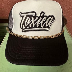 May include: Black and white trucker hat with a gold chain trim. The hat has a white mesh back and a black front with the word "Toxica" embroidered in white.