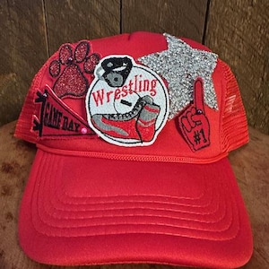 May include: Red mesh trucker hat with various patches including a paw print, a wrestling shoe, a star, and a number one finger. The patches are all glittery and colorful.