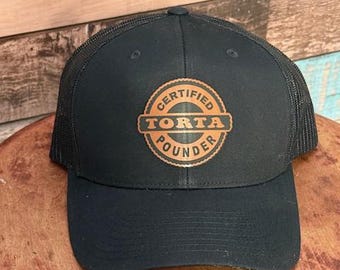 May include: Black trucker hat with a curved brim and mesh back. The hat features a circular patch with the words "CERTIFIED TORTA POUNDER" in a copper-colored font. The hat is sitting on a wooden surface.