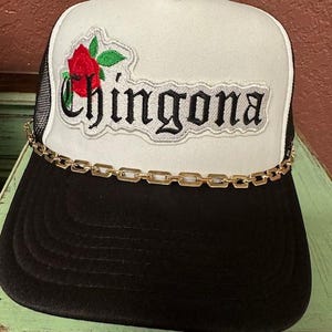 May include: A white and black trucker hat with a gold chain trim. The front of the hat features a white patch with black text that reads "Chingona" and a red rose with green leaves.
