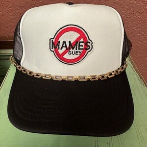 May include: A black and white trucker hat with a red circle and white text that says "MAMES GUEY" with a gold chain around the brim.