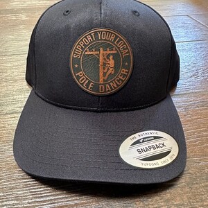 May include: Black baseball cap with a flat brim. The cap features a circular patch with the text "SUPPORT YOUR LOCAL POLE DANCER" around an image of a lineman on a utility pole. The tag on the brim reads "SNAPBACK".