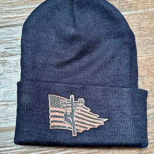 May include: Black knit beanie with a brown leather patch featuring an American flag with a lineman climbing a utility pole.
