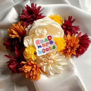 May include: A white sticker with the text "Read More Books" in colorful letters with a flower design. The sticker is placed on a bouquet of white, yellow, orange, and burgundy flowers.