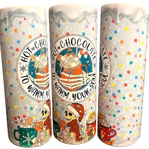 May include: Three white tumblers with a glittery background and a winter-themed design. The design features skeletons holding mugs of hot chocolate with the text "Hot Chocolate to Warm Your Soul".
