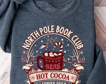 May include: A dark gray sweatshirt with a festive design. The graphic features a mug of hot cocoa, reindeer, and the text "North Pole Book Club." The design also includes the words "Read," "Hot Cocoa Served Daily," and "Warm Up Here Open All Winter."
