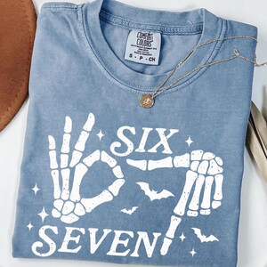 May include: A blue Comfort Colors t-shirt with a skeleton hand graphic that spells out "Six Seven". The shirt has a crew neck and short sleeves. A gold necklace with a pendant is draped across the shirt.
