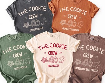 May include: Five t-shirts in various colors, each featuring the text "THE COOKIE CREW EST. 2026" with cookie-themed graphics and job titles like "DOUGH ROLLER" and "HEAD BAKER". The shirts are in shades of gray, orange, green, cream, and brown.