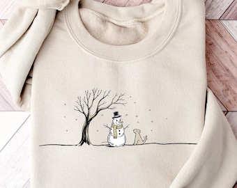 May include: Beige sweatshirt featuring a black and white illustration of a snowman wearing a top hat and scarf, standing under a bare tree with a dog. The design is centered on the front of the folded sweatshirt.