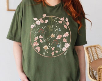 May include: A green t-shirt with a circular floral print. The print features pink and white flowers and leaves in a circle on a green background.