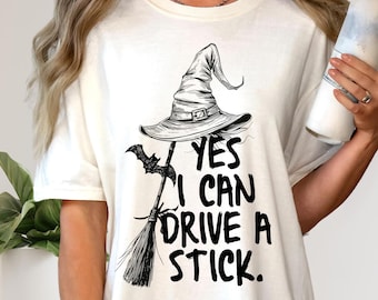 May include: Cream-colored t-shirt with a black graphic of a witch's hat, bat, and broom. The text "YES I CAN DRIVE A STICK" is printed below the hat. The design is perfect for Halloween.