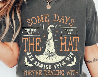 May include: Dark gray t-shirt with orange and white text and a witch hat graphic. The text reads "SOME DAYS YOU JUST HAVE TO PUT ON THE HAT AND REMIND THEM WHO THEY'RE DEALING WITH."