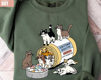 May include: A green sweatshirt with a graphic of a pill bottle labeled "Serotonin Booster" with a group of cartoon cats spilling out of it. The text on the pill bottle reads "Side Effect: Cute Aggression".