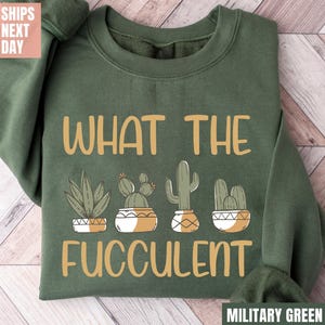 May include: Military green sweatshirt with the text "WHAT THE FUCCULENT" and illustrations of cacti and succulents in pots. The sweatshirt is a crew neck style.