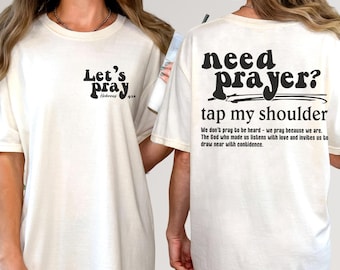May include: Cream-colored t-shirt with black text. The front reads "Let's Pray" with a Bible verse reference. The back says "Need Prayer? Tap my shoulder" with a supporting quote. A casual, faith-based apparel item.