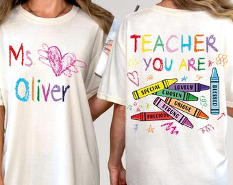 May include: Cream-colored t-shirt with colorful crayon-style text and graphics. The front features "Ms. Oliver" and a heart design. The back says "Teacher You Are" with crayon illustrations and words like "Special" and "Blessed".