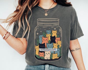May include: Dark grey t-shirt featuring a whimsical illustration of colorful cats inside a jar, with the word "ANTIDEPRESSANTS" on a banner. The design includes various cat colors and leafy accents, creating a playful and comforting aesthetic.