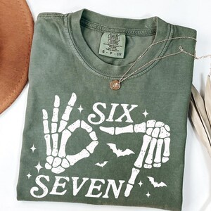 May include: Olive green t-shirt with a white skeleton hand graphic spelling out "SIX SEVEN" with bats and stars. A gold necklace with a pendant rests on the shirt. The shirt is folded and has a Comfort Colors tag.