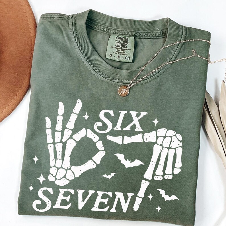 May include: Olive green t-shirt with a white skeleton hand graphic spelling out "SIX SEVEN" with bats and stars. A gold necklace with a pendant rests on the shirt. The shirt is folded and has a Comfort Colors tag.