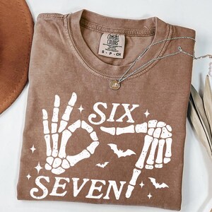 May include: A brown Comfort Colors t-shirt with a white skeleton hand graphic forming the number six and seven. The shirt has a crew neck and short sleeves. A silver necklace with a small pendant is draped across the shirt.