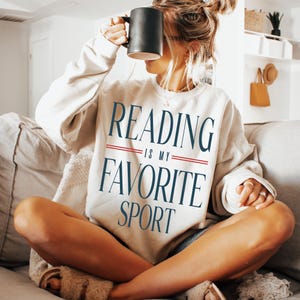 May include: A cream-coloured sweatshirt with the text "READING IS MY FAVORITE SPORT" in navy blue lettering and a red stripe. The person is holding a black mug.