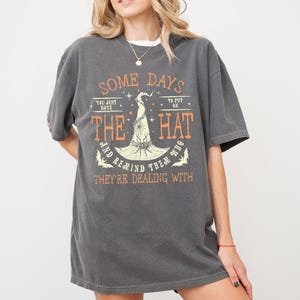 May include: Dark gray t-shirt with the text "SOME DAYS YOU JUST BAYE THE HAT AND REMIND THEM WHO THEY'RE DEALING WITH." The graphic features a witch's hat and bats in orange. The shirt has short sleeves.