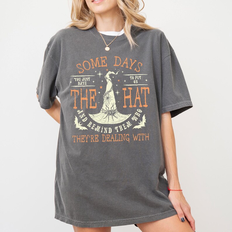 May include: Dark gray t-shirt with the text "SOME DAYS YOU JUST BAYE THE HAT AND REMIND THEM WHO THEY'RE DEALING WITH." The graphic features a witch's hat and bats in orange. The shirt has short sleeves.