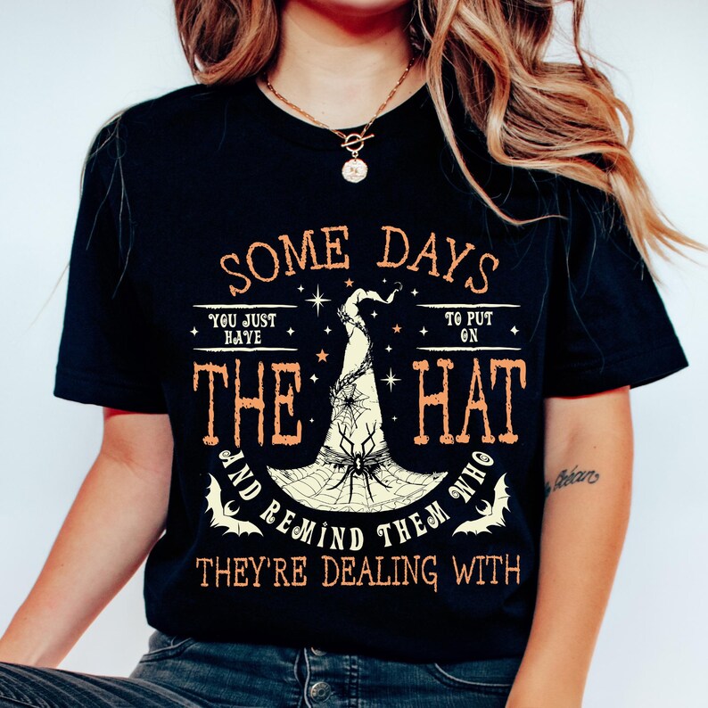 May include: Black t-shirt with orange text and a witch hat graphic. The text reads "SOME DAYS YOU JUST HAVE TO PUT ON THE HAT AND REMIND THEM WHO THEY'RE DEALING WITH."