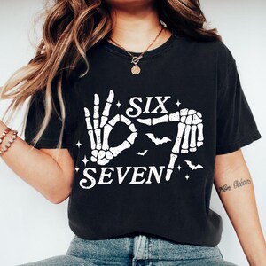May include: Black t-shirt with a white skeleton hand design spelling out "SIX SEVEN" with bats and stars. The shirt is a casual, comfortable style.