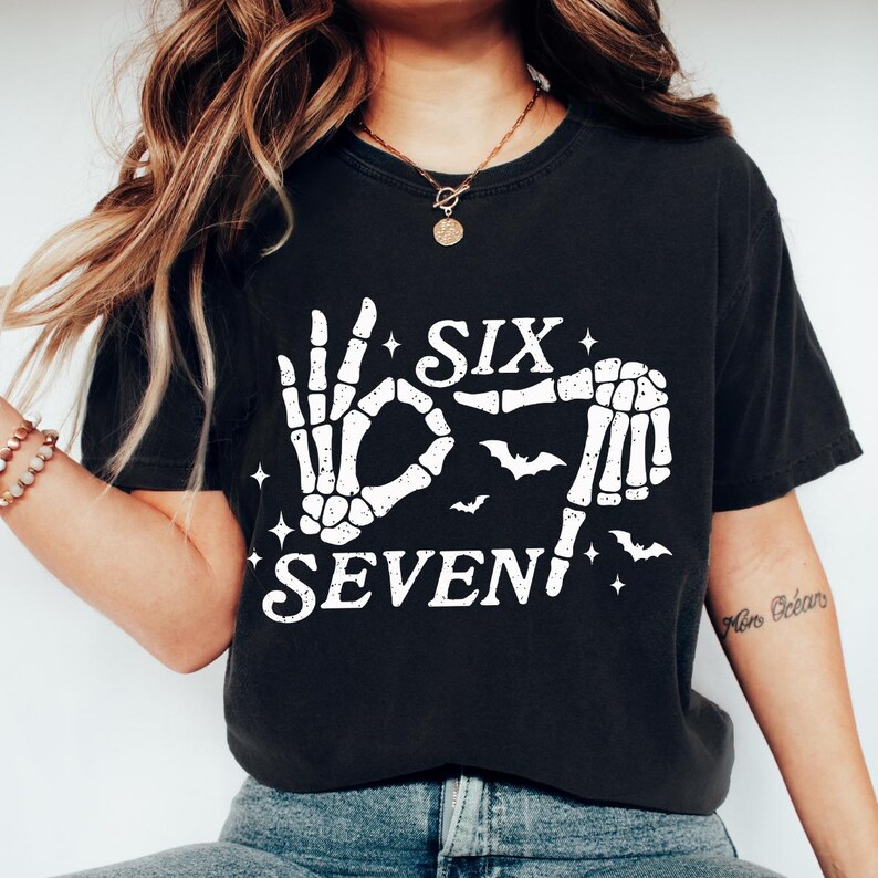 May include: Black t-shirt with a white skeleton hand design spelling out "SIX SEVEN" with bats and stars. The shirt is a casual, comfortable style.