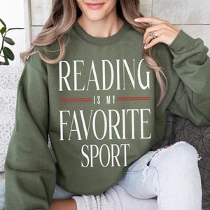May include: An olive green sweatshirt with the white text "READING IS MY FAVORITE SPORT". The text is centered, with two red lines separating the phrases. The sweatshirt is a casual style.