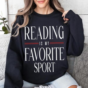 May include: Black sweatshirt featuring the white text "READING IS MY FAVORITE SPORT", separated by two red lines. The sweatshirt has a crew neck design.