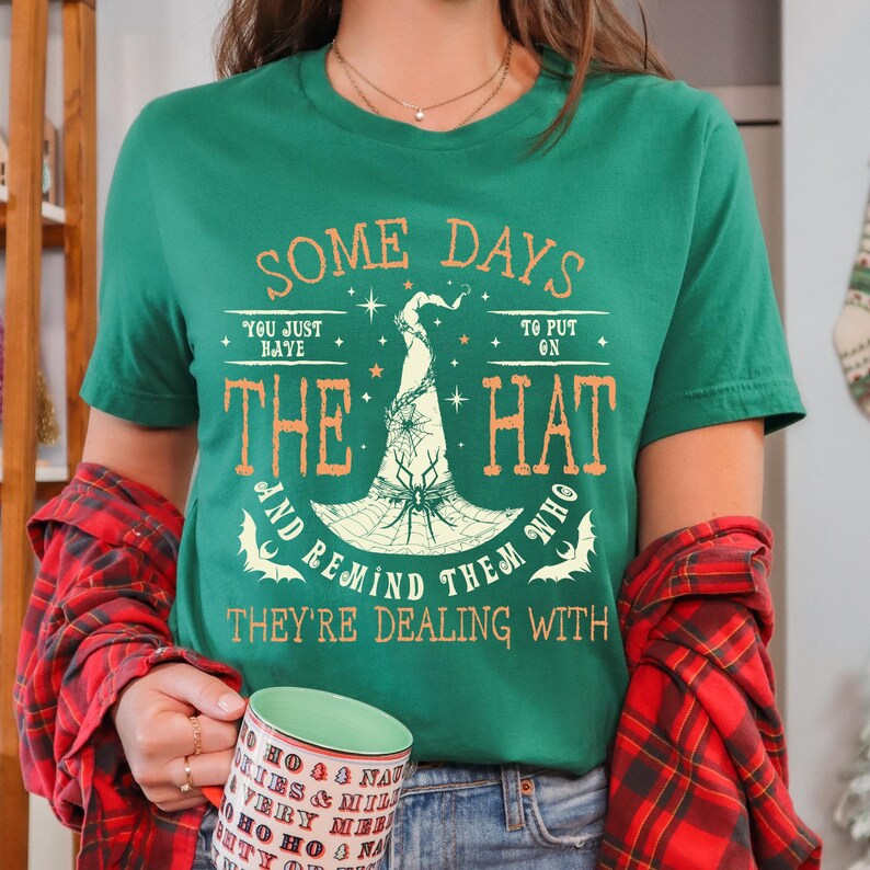 May include: A green t-shirt with the text "SOME DAYS YOU JUST HAVE TO PUT ON THE HAT AND REMIND THEM WHO THEY'RE DEALING WITH." The design includes a witch hat with a spider web and bats. The person is holding a mug.