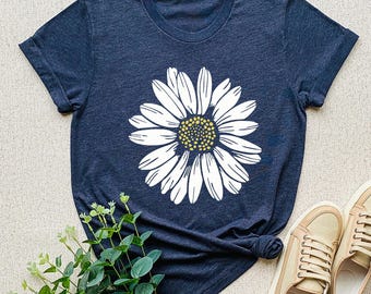 May include: Navy blue t-shirt featuring a large white daisy graphic with a yellow center. The shirt has short sleeves and a crew neck. The shirt is styled with tan sneakers and greenery.