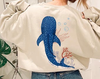May include: A cream-colored sweatshirt featuring a large blue whale shark design on the back. The design includes blue bubbles, red coral, and seaweed. The sweatshirt is a relaxed fit.