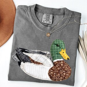 May include: A charcoal gray short-sleeve t-shirt featuring a large mallard duck appliqué made from various fabric patches. The duck has a green gingham head, a yellow polka-dot beak, and a brown floral chest. The shirt label reads Comfort Colors.