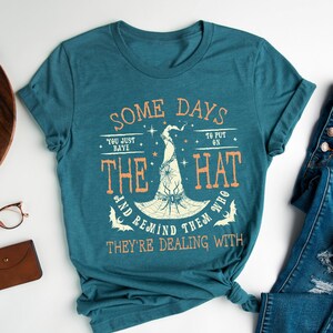 May include: Teal t-shirt with the text "SOME DAYS YOU JUST HAVE TO PUT ON THE HAT AND REMIND THEM WHO THEY'RE DEALING WITH." The design features a witch hat with a spider web and bats. The shirt is folded and knotted at the bottom.