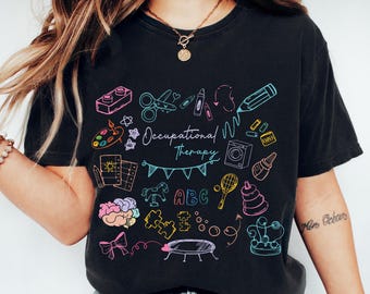 May include: Black t-shirt with a colorful graphic design featuring the words "Occupational Therapy" and various playful illustrations. The design includes a pencil, scissors, paint palette, and building blocks.