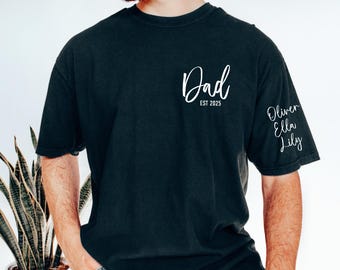 May include: Black t-shirt with white script text that reads "Dad EST 2025" on the chest. The sleeve features the names "Oliver, Ella, Lily" in white script. The shirt is a casual, comfortable style.