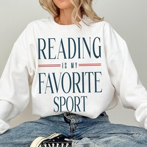 May include: White sweatshirt featuring the navy blue text "READING IS MY FAVORITE SPORT", separated by red lines. The sweatshirt has a crew neck design.