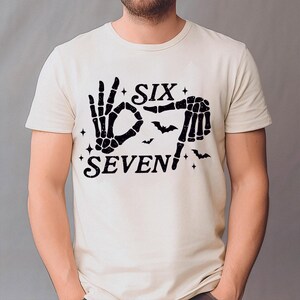 May include: Cream-colored t-shirt with a black skeleton hand graphic forming the numbers six and seven. The word "SIX" is above the hand gesture, and "SEVEN" is below. Small bats and stars complete the design.