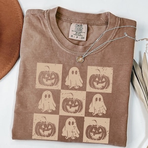 May include: A brown Comfort Colors t-shirt with a checkerboard pattern of jack-o'-lanterns and ghosts. The shirt has a crew neck and short sleeves. A silver necklace with a small pendant is draped across the shirt.