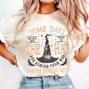 May include: Cream-colored t-shirt with the text "SOME DAYS YOU JUST HAVE TO PUT ON THE HAT AND REMIND THEM WHO THEY'RE DEALING WITH." The design features a black witch hat with a spiderweb and bats, in orange and black.
