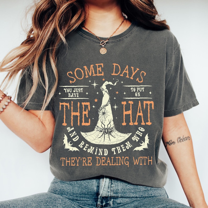 May include: Dark gray t-shirt with orange and white text and a witch hat graphic. The text reads "SOME DAYS YOU JUST HAVE TO PUT ON THE HAT AND REMIND THEM WHO THEY'RE DEALING WITH."