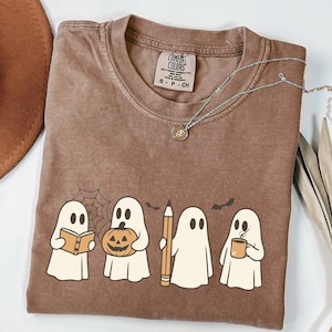 May include: Brown Comfort Colors t-shirt featuring a graphic of four cartoon ghosts. One ghost holds a book, another a pumpkin, a third a pencil, and the last a mug. A silver necklace with a pendant is also visible.