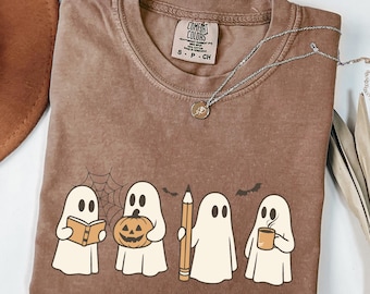 Può includere: T-shirt Comfort Colors marrone con una grafica di quattro fantasmi dei cartoni animati. Un fantasma tiene in mano un libro, un altro una zucca, un terzo una matita e l'ultimo una tazza. È visibile anche una collana d'argento con un ciondolo.