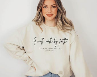 May include: Cream-colored sweatshirt with the text "I will walk by faith" in a black script font. Below the main text is the phrase "EVEN WHEN I CANNOT SEE" and the reference "2 CORINTHIANS 5:7". The sweatshirt is a casual style.